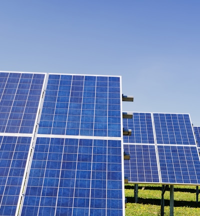A field of solar panels arranged in rows under a clear blue sky, capturing sunlight to convert into energy. The panels are mounted on metal structures above a grassy area.