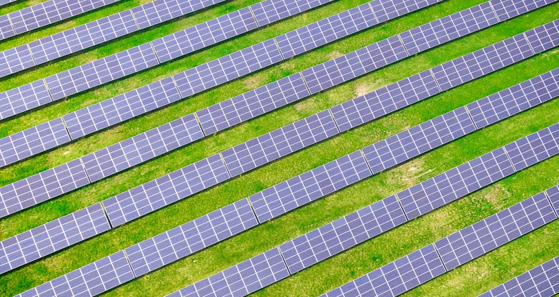 An array of solar panels arranged in parallel rows on a lush green field, capturing sunlight for energy conversion. The solar panels create a pattern of alternating blue and green stripes.
