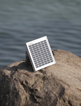 A solar panel is positioned on a large rock near a body of water, suggesting energy harnessing in a natural setting.