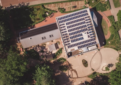 An aerial view of a building with a large array of solar panels on its roof. The structure is surrounded by greenery, pathways, and some smaller structures or landscaped areas. The environment appears to be a mix of natural and man-made elements, suggesting a focus on sustainability.