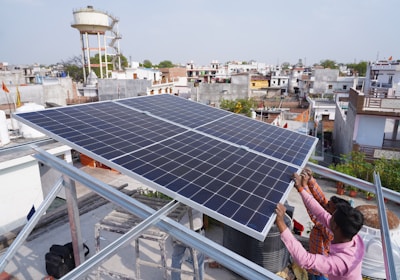Two people are installing a large solar panel on a rooftop in an urban area with numerous buildings and a water tower in the background. The scene suggests an effort to set up renewable energy resources.