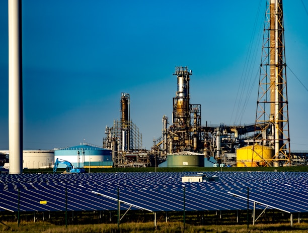 A landscape featuring a large solar panel array in the foreground, with a modern wind turbine to the left. Behind the renewable energy installations, an industrial facility with multiple chimneys and storage tanks is visible under a clear blue sky.
